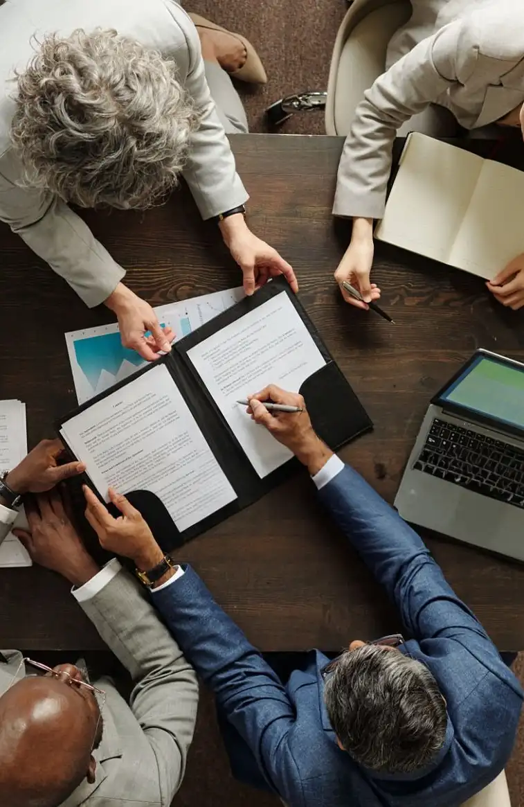 Reunião vista de cima, com documentos, notebook e pessoas à mesa.